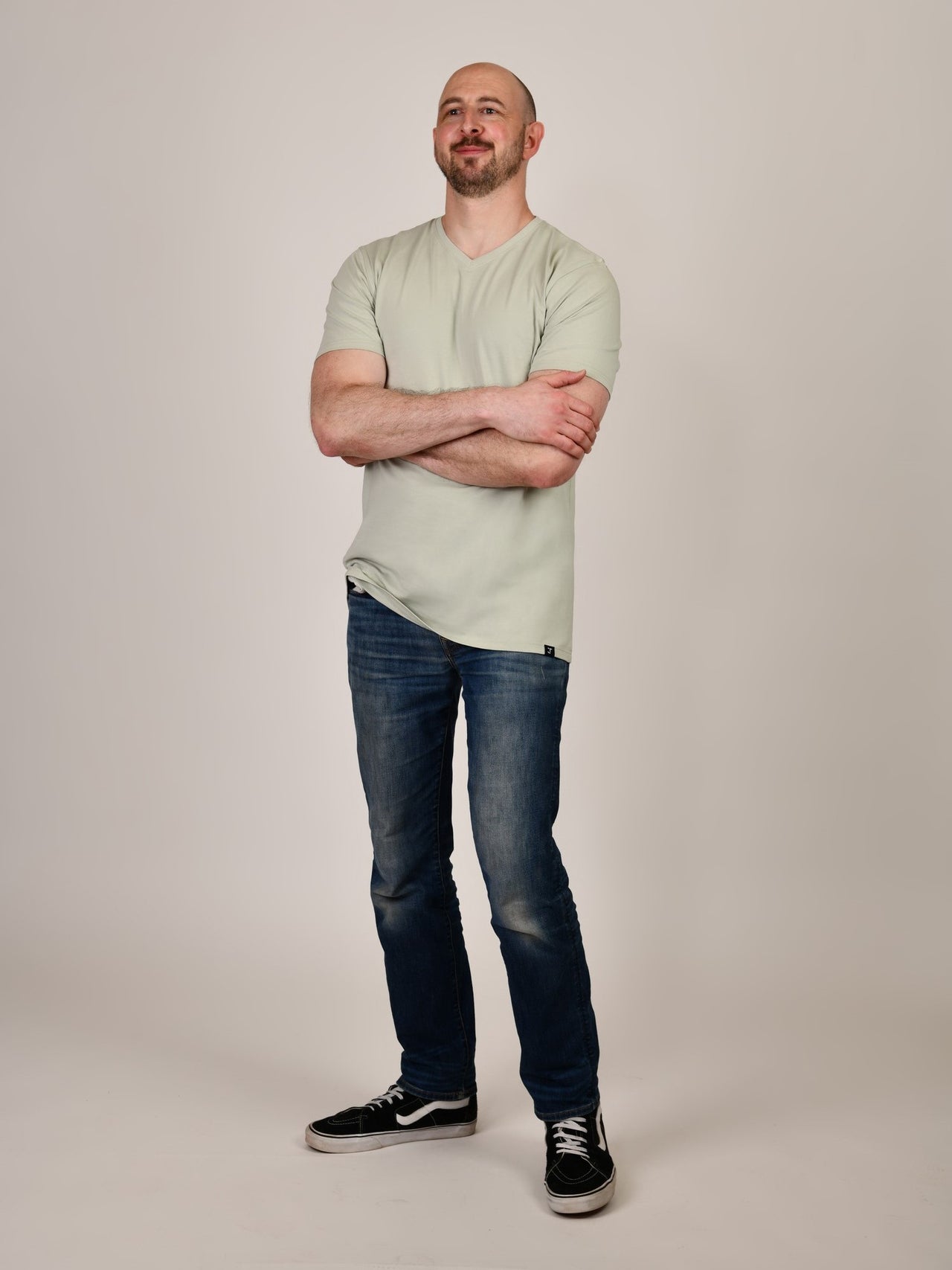 A head to toe shot of a tall and slim guy in the studio, hands folded and wearing a sage green XL tall slim v-neck t-shirt.
