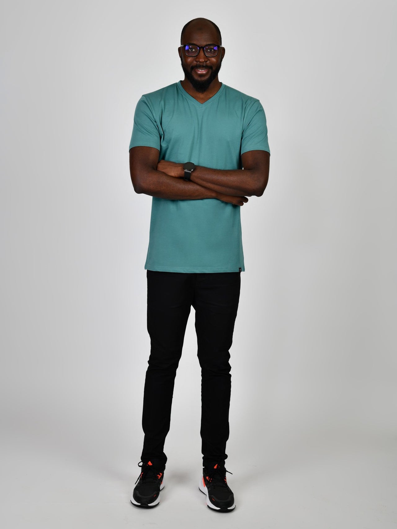 A head to toe shot of a tall and slim guy in the studio, hands folded and wearing a teal L tall slim v-neck t-shirt.