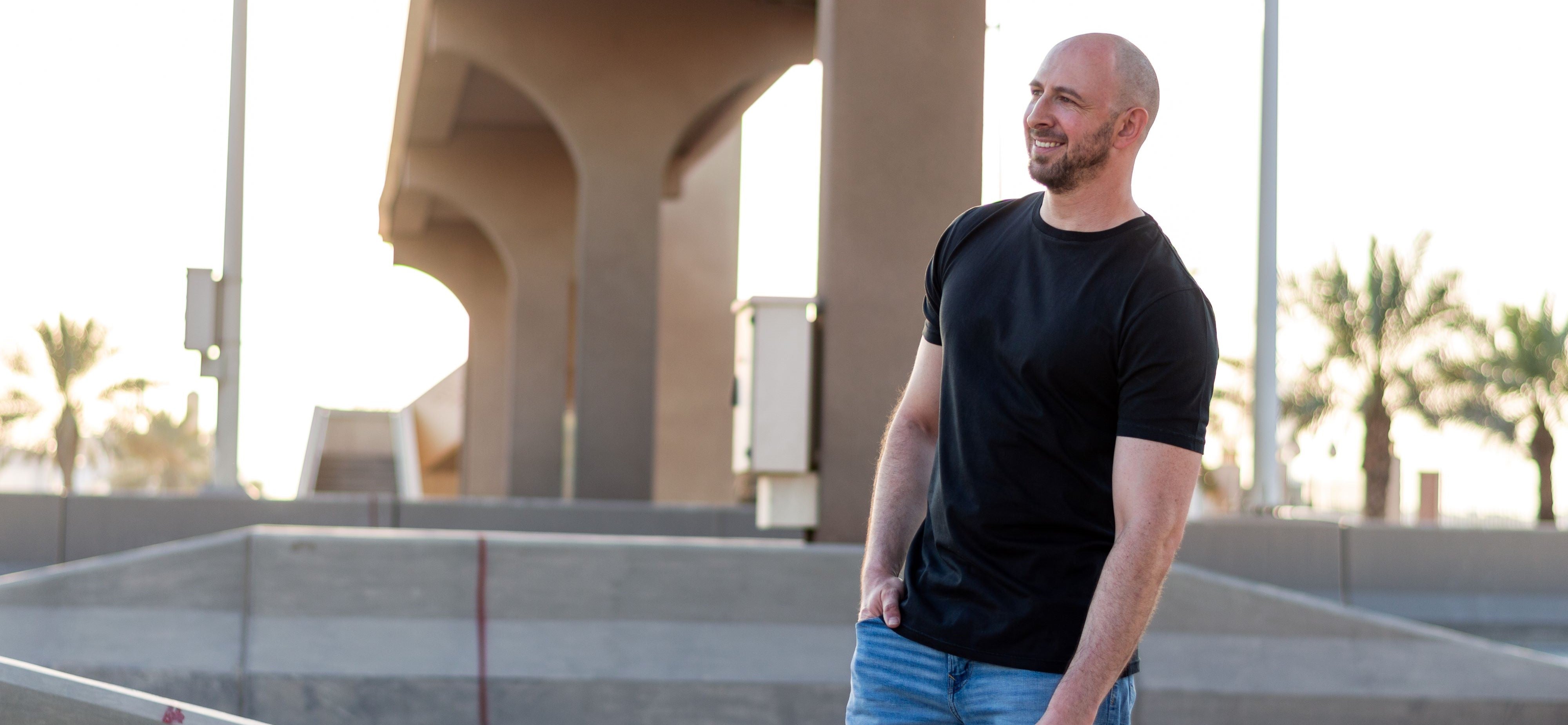 A tall slim guy smiling under a bridge and wearing a black XL tall extra long t-shirt.