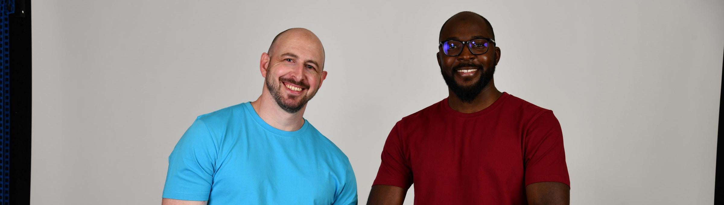 Two tall slim guys smiling in a studio and wearing cyan and maroon crew neck tall t-shirts.