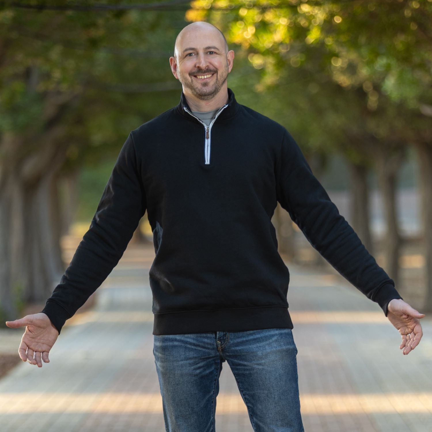 A tall slim man standing on a path in a park and wearing an extra long tall quarter zip black sweatshirt.