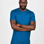 An upper body shot of a tall slim guy wearing an extra long large tall azure t-shirt, hands folded and looking serious.