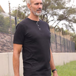 A tall and slim man standing outside. The model is wearing an extra long slim black henley shirt. The tall black henley shirt features a 3" longer body, 100% organic cotton, and is soft & preshrunk. The black henley shirt is ideal for tall slim men 6'2"+.