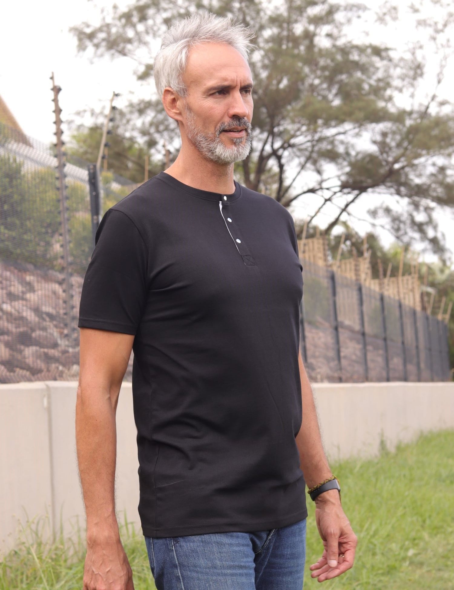 A tall and slim man standing outside. The model is wearing an extra long slim black henley shirt. The tall black henley shirt features a 3" longer body, 100% organic cotton, and is soft & preshrunk. The black henley shirt is ideal for tall slim men 6'2"+.