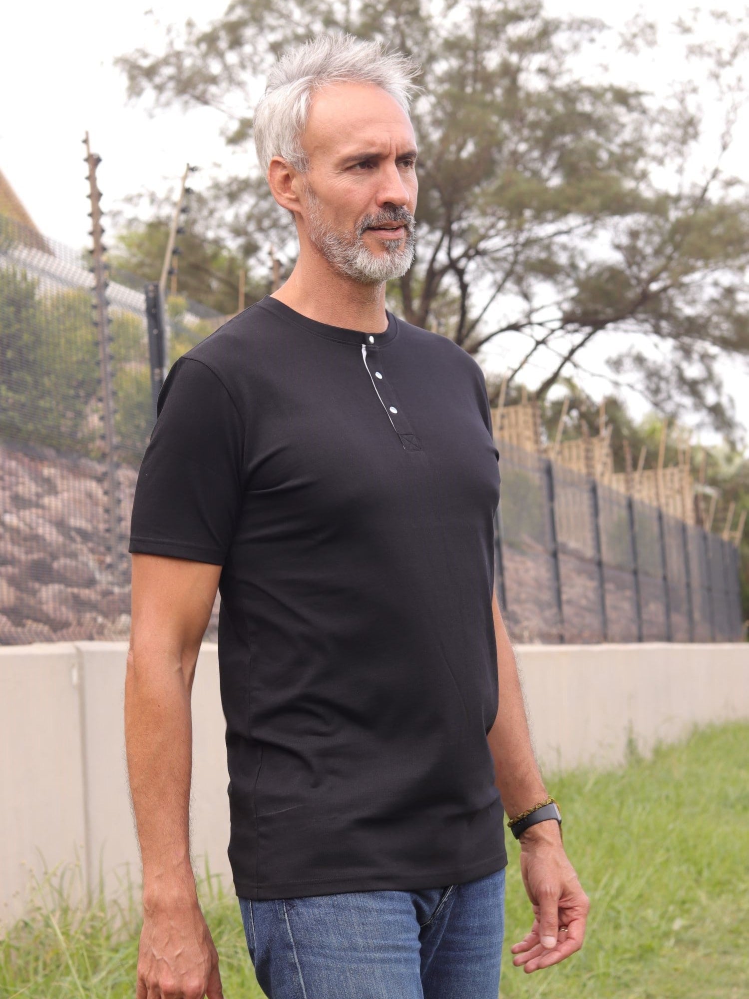 A tall and slim man standing outside. The model is wearing an extra long slim black henley shirt. The tall black henley shirt features a 3" longer body, 100% organic cotton, and is soft & preshrunk. The black henley shirt is ideal for tall slim men 6'2"+.