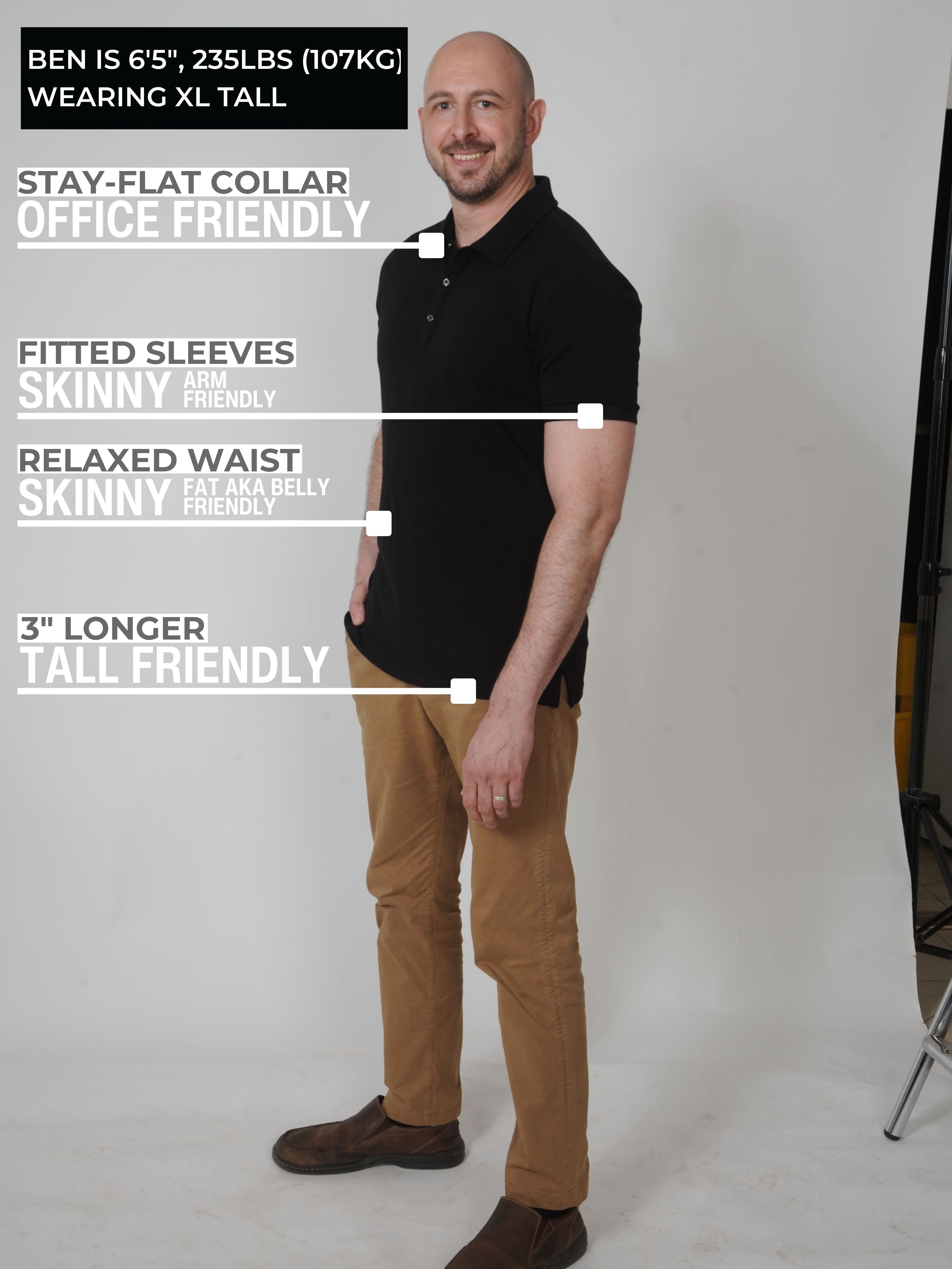 A head to toe shot of a tall slim man in a studio wearing a black XL tall polo shirt, smiling and with hand in pocket.