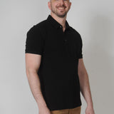 A tall and slim man in the studio standing in front of a light background. The smiling model is wearing an extra long slim black polo shirt in a size XL. The tall black polo shirt features a 3" longer body, 100% organic cotton, and is soft & preshrunk. The black polo shirt is ideal for tall slim men 6'2"+.