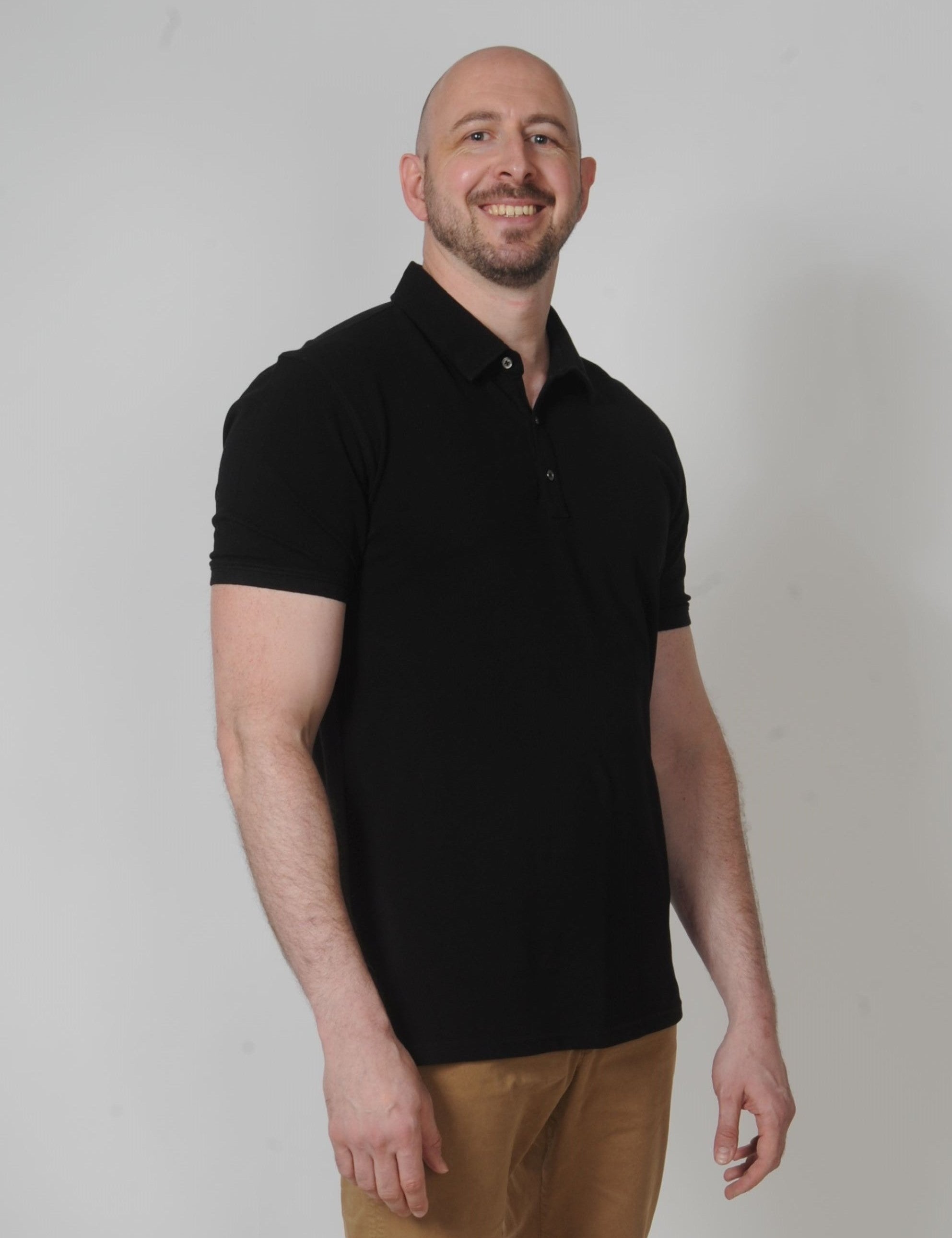 A tall and slim man in the studio standing in front of a light background. The smiling model is wearing an extra long slim black polo shirt in a size XL. The tall black polo shirt features a 3" longer body, 100% organic cotton, and is soft & preshrunk. The black polo shirt is ideal for tall slim men 6'2"+.