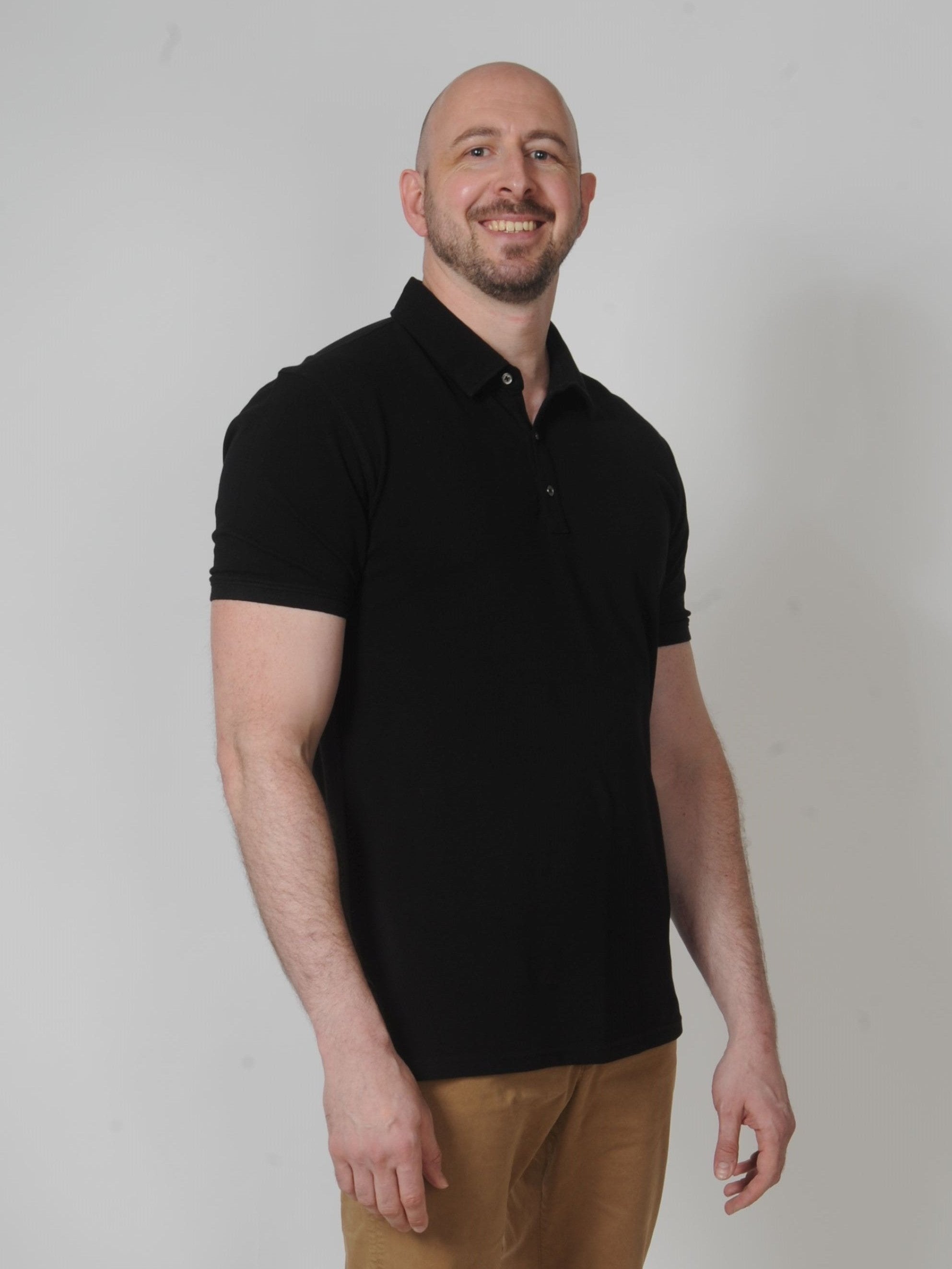 A tall and slim man in the studio standing in front of a light background. The smiling model is wearing an extra long slim black polo shirt in a size XL. The tall black polo shirt features a 3" longer body, 100% organic cotton, and is soft & preshrunk. The black polo shirt is ideal for tall slim men 6'2"+.