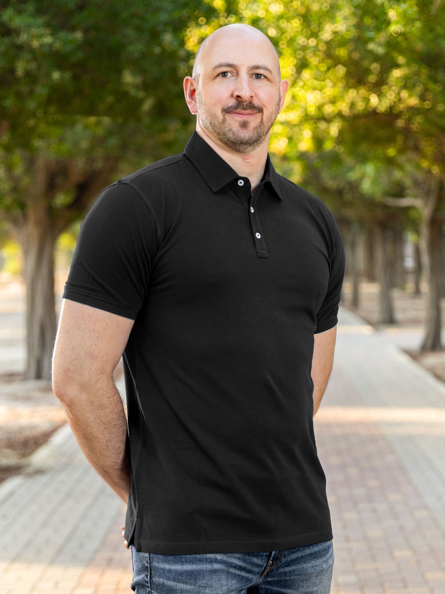 A tall and athletic man in a park standing on a path between trees. The smiling model is wearing an extra long slim black polo shirt in a size XL. The tall black polo shirt features a 3" longer body, 100% organic cotton, and is soft & preshrunk. The black polo shirt is ideal for tall slim men 6'2"+.