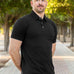 A tall and athletic man in a park standing on a path between trees. The smiling model is wearing an extra long slim black polo shirt in a size XL. The tall black polo shirt features a 3" longer body, 100% organic cotton, and is soft & preshrunk. The black polo shirt is ideal for tall slim men 6'2"+.