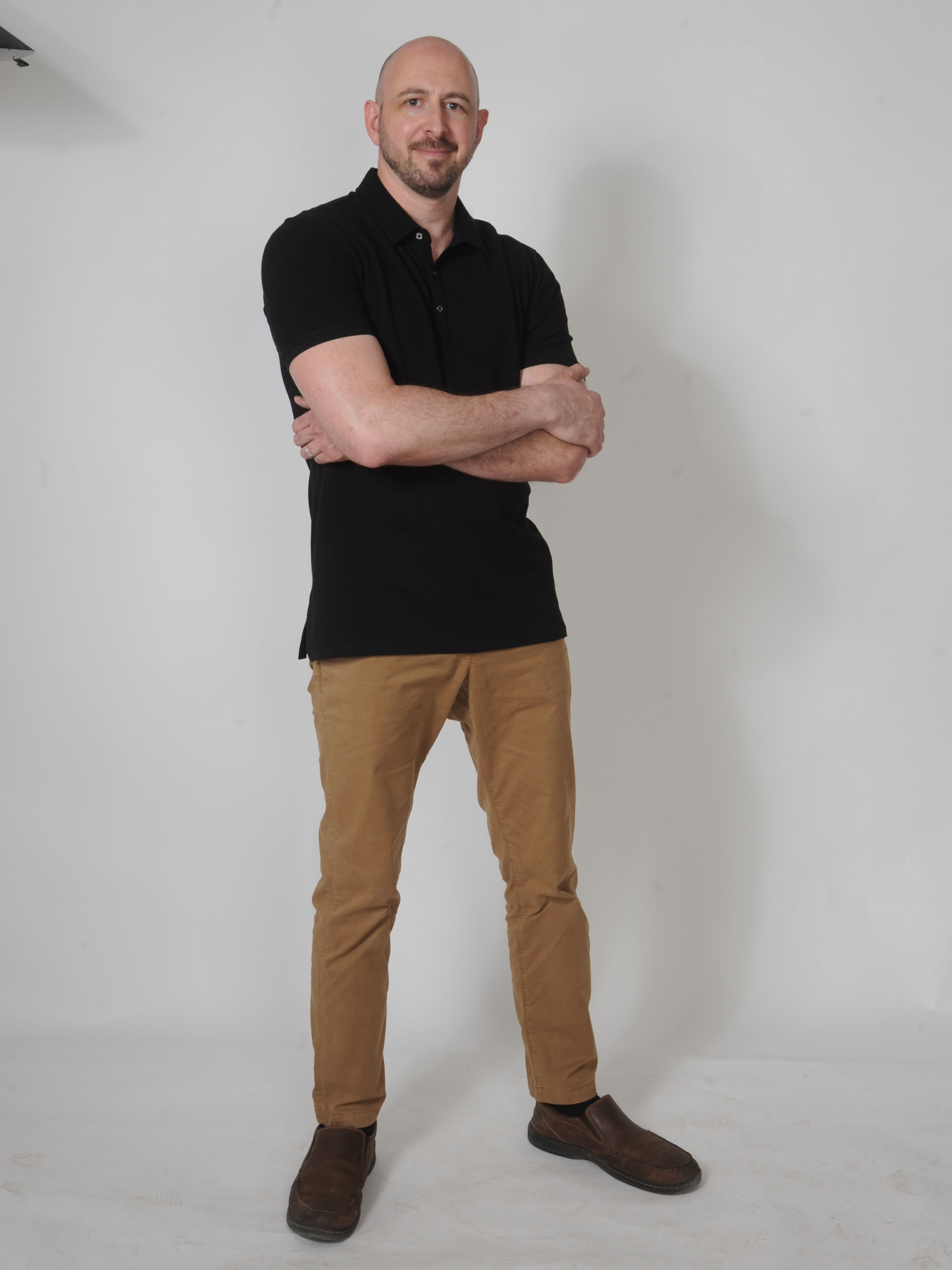 A head to toe shot of a tall skinny guy wearing an XL tall black pique polo shirt in a studio with hands folded.