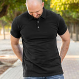 A tall athletic guy in a park with his hands behind his back, looking down at his waist and wearing a black XL tall polo shirt.