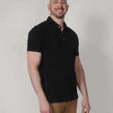 A tall and slim man in the studio standing in front of a light background. The smiling model is wearing an extra long slim black polo shirt in a size XL. The tall black polo shirt features a 3" longer body, 100% organic cotton, and is soft & preshrunk. The black polo shirt is ideal for tall slim men 6'2"+.