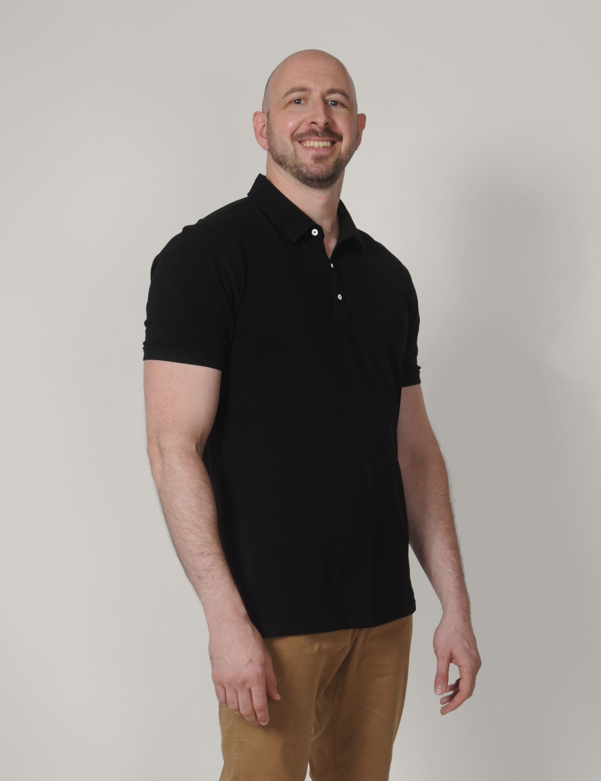 A tall and slim man in the studio standing in front of a light background. The smiling model is wearing an extra long slim black polo shirt in a size XL. The tall black polo shirt features a 3" longer body, 100% organic cotton, and is soft & preshrunk. The black polo shirt is ideal for tall slim men 6'2"+.