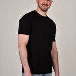 A tall and slim man in the studio standing in front of a light background. The smiling model is wearing an extra long slim black t-shirt in a size XL. The tall black t-shirt features a 3" longer body, 100% organic cotton, and is soft & preshrunk. The black t-shirt is ideal for tall slim men 6'2"+.