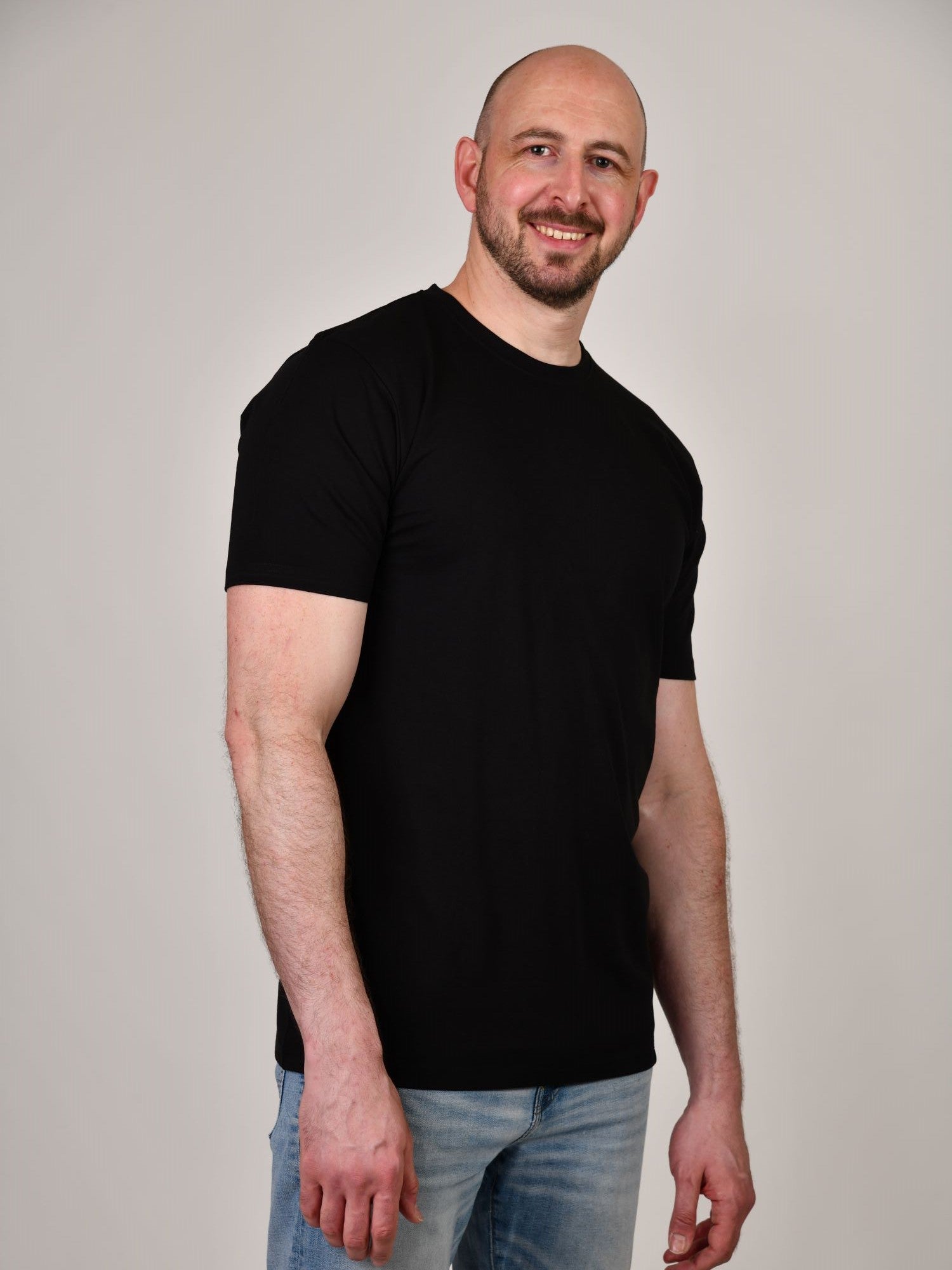 A tall and slim man in the studio standing in front of a light background. The smiling model is wearing an extra long slim black t-shirt in a size XL. The tall black t-shirt features a 3" longer body, 100% organic cotton, and is soft & preshrunk. The black t-shirt is ideal for tall slim men 6'2"+.