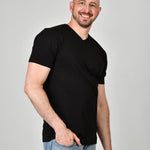 A tall and slim man in the studio standing in front of a light background with one hand in his pocket. The smiling model is wearing an extra long slim black v-neck t-shirt in a size XL. The tall black v-neck t-shirt features a 3" longer body, 100% organic cotton, and is soft & preshrunk. The black v-neck t-shirt is ideal for tall slim men 6'2"+.