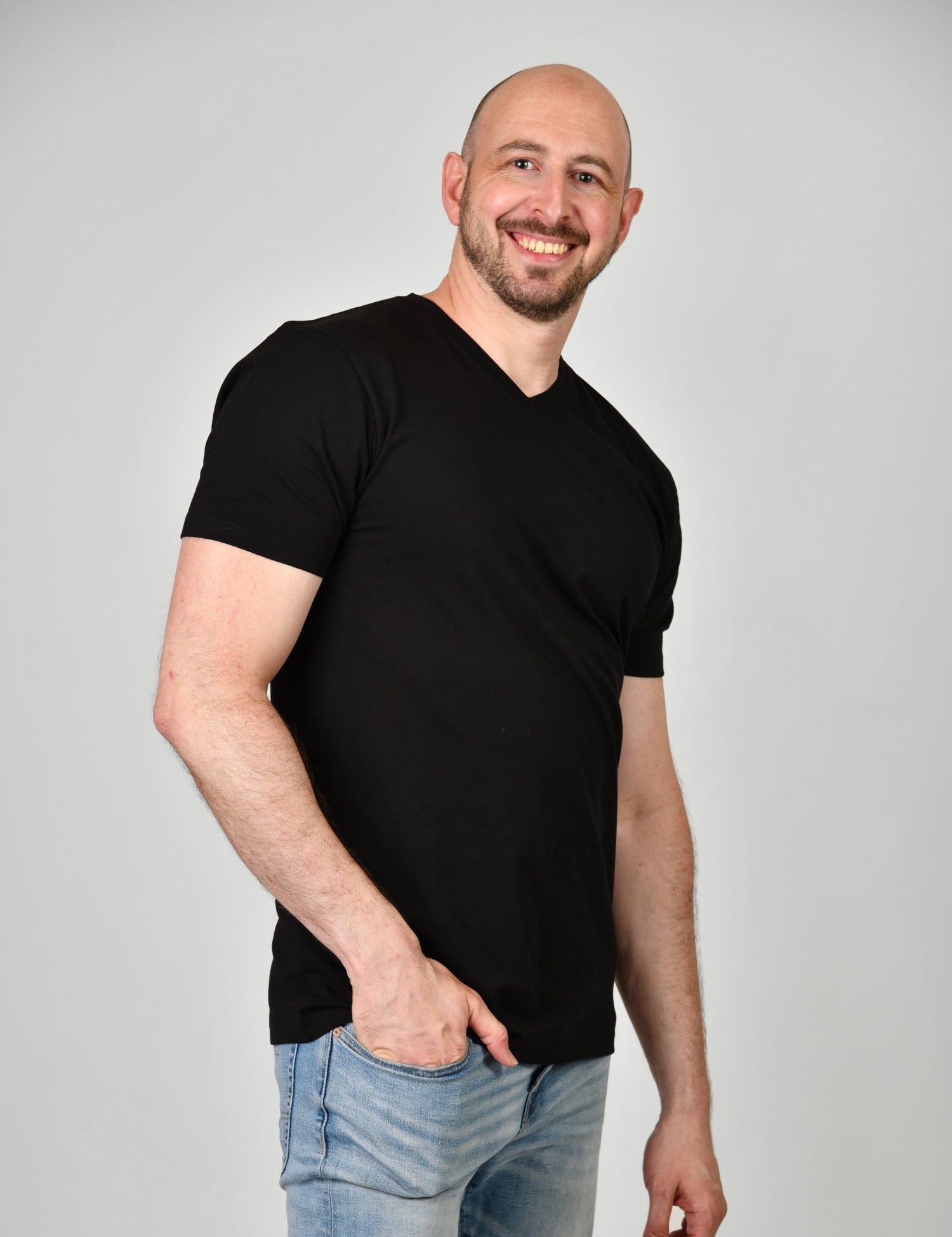 A tall and slim man in the studio standing in front of a light background with one hand in his pocket. The smiling model is wearing an extra long slim black v-neck t-shirt in a size XL. The tall black v-neck t-shirt features a 3" longer body, 100% organic cotton, and is soft & preshrunk. The black v-neck t-shirt is ideal for tall slim men 6'2"+.