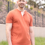 A tall and slim man standing outside. The model is wearing an extra long slim brown henley shirt. The tall brown henley shirt features a 3" longer body, 100% organic cotton, and is soft & preshrunk. The brown henley shirt is ideal for tall slim men 6'2"+.