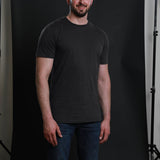 A tall and slim man in the studio standing in front of a dark background. The smiling model is wearing an extra long slim charcoal t-shirt in size a XL. The tall charcoal t-shirt features a 3" longer body, 100% organic cotton, and is soft & preshrunk. The charcoal t-shirt is ideal for tall slim men 6'2"+.