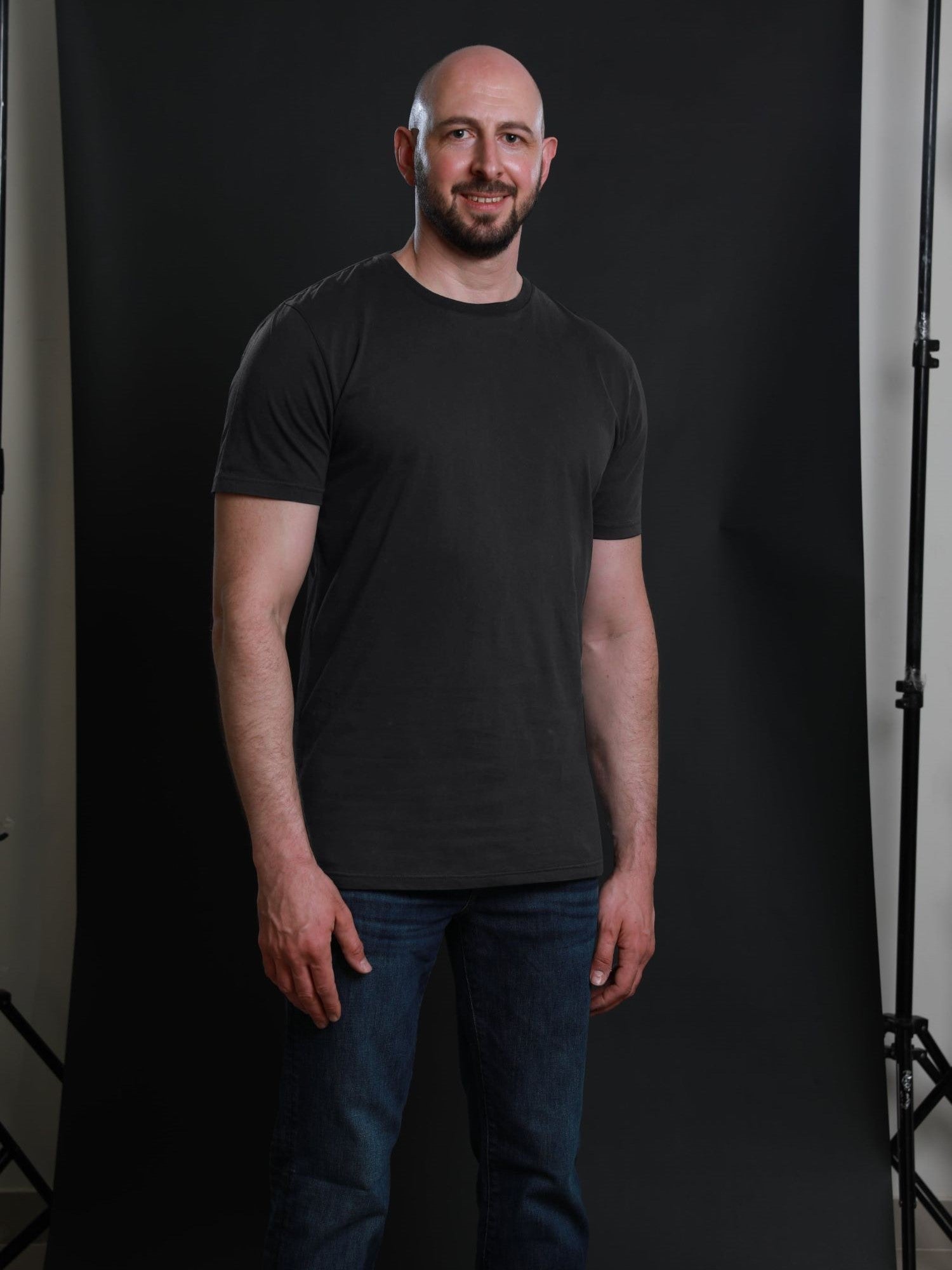 A tall and slim man in the studio standing in front of a dark background. The smiling model is wearing an extra long slim charcoal t-shirt in size a XL. The tall charcoal t-shirt features a 3" longer body, 100% organic cotton, and is soft & preshrunk. The charcoal t-shirt is ideal for tall slim men 6'2"+.