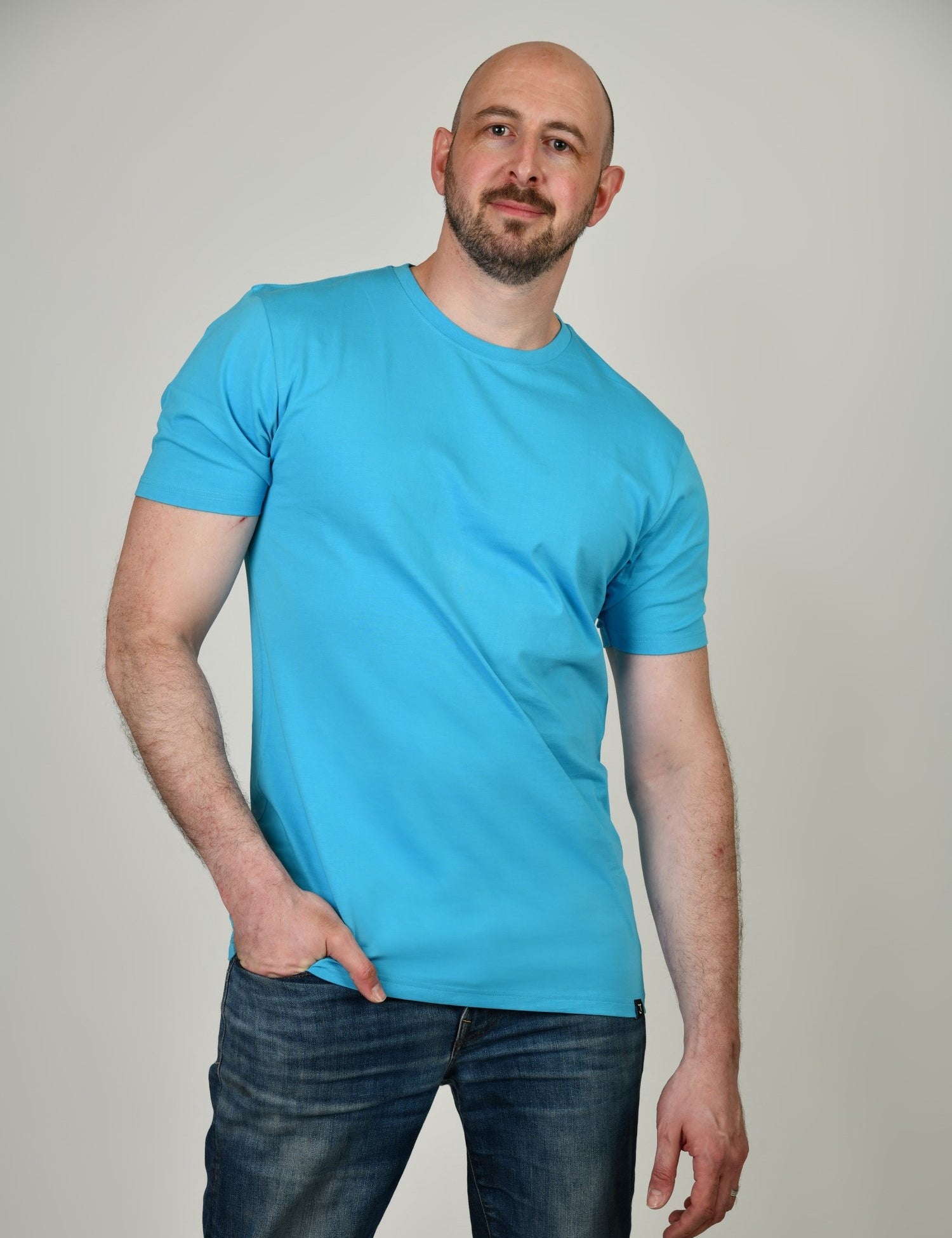 A tall and slim man in the studio standing in front of a light background with one hand in his pocket. The model is wearing an extra long slim cyan blue t-shirt in a size XL. The cyan blue t-shirt features a 3" longer body, 100% organic cotton, and is soft & preshrunk. The cyan blue t-shirt is ideal for tall slim men 6'2"+.
