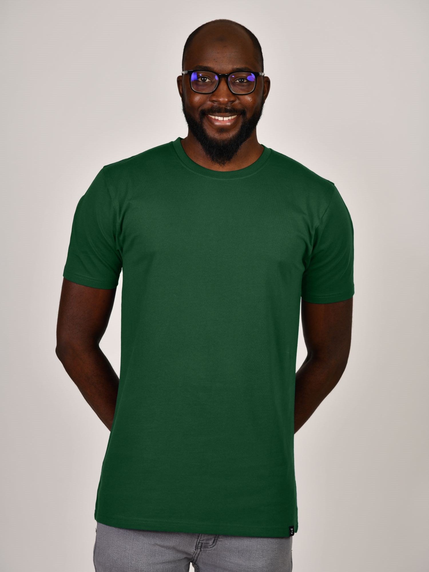 A tall and slim guy smiling in the studio, hands behind back and wearing a dark green L tall slim t-shirt.