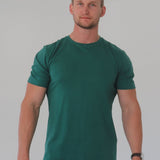 A tall and broad man in the studio standing in front of a light background. The model is wearing an extra long slim dark green t-shirt in a size 2XL. The dark green t-shirt features a 3" longer body, 100% organic cotton, and is soft & preshrunk. The dark green t-shirt is ideal for tall slim men 6'2"+.