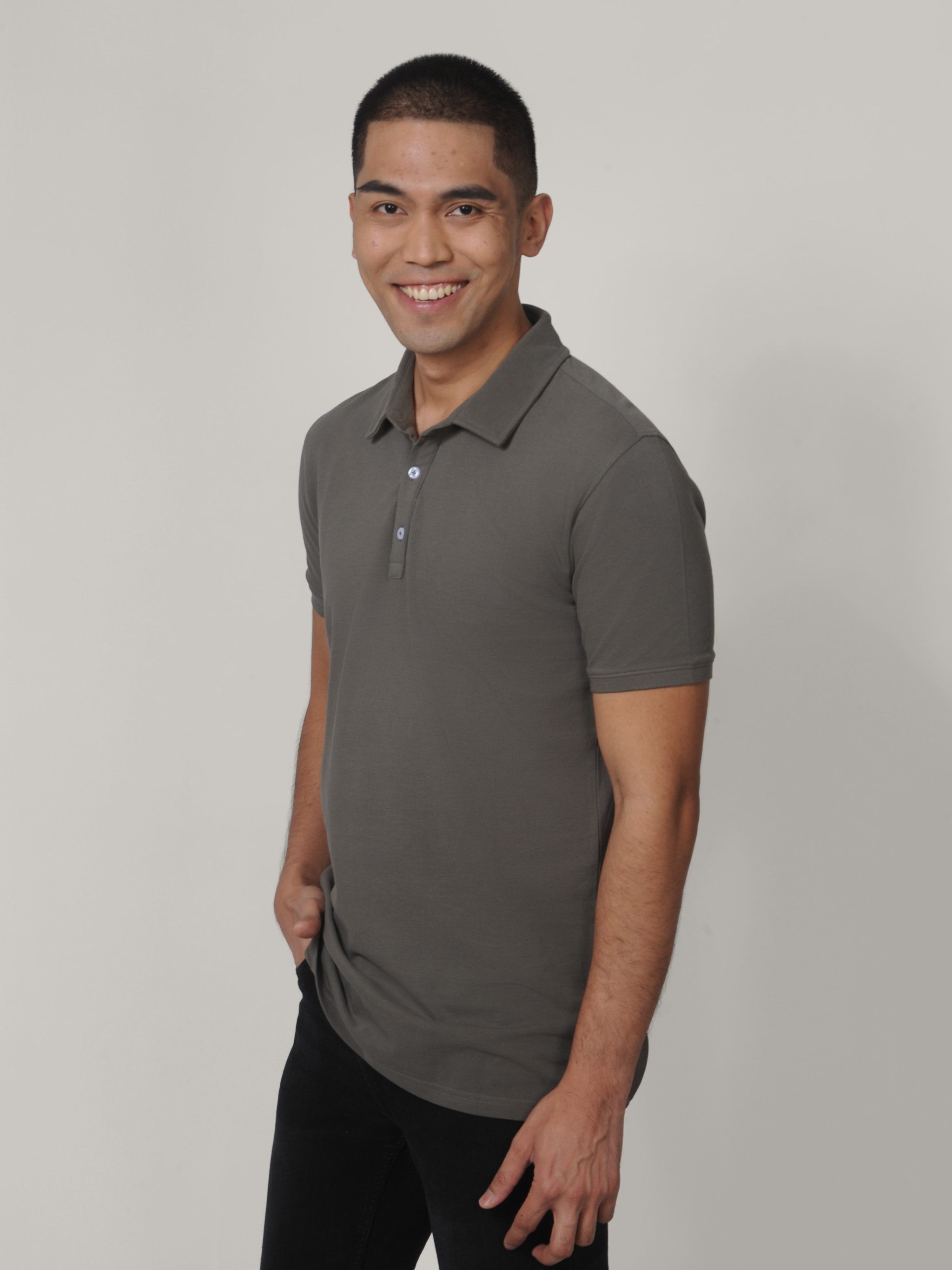 A tall and slim man in the studio standing in front of a light background. The smiling model is wearing an extra long slim dark grey polo shirt in a size medium. The tall dark grey polo shirt features a 3" longer body, 100% organic cotton, and is soft & preshrunk. The dark grey polo shirt is ideal for tall slim men 6'2"+.