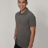 A tall and slim man in the studio standing in front of a light background. The smiling model is wearing an extra long slim dark grey polo shirt in a size medium. The tall dark grey polo shirt features a 3" longer body, 100% organic cotton, and is soft & preshrunk. The dark grey polo shirt is ideal for tall slim men 6'2"+.