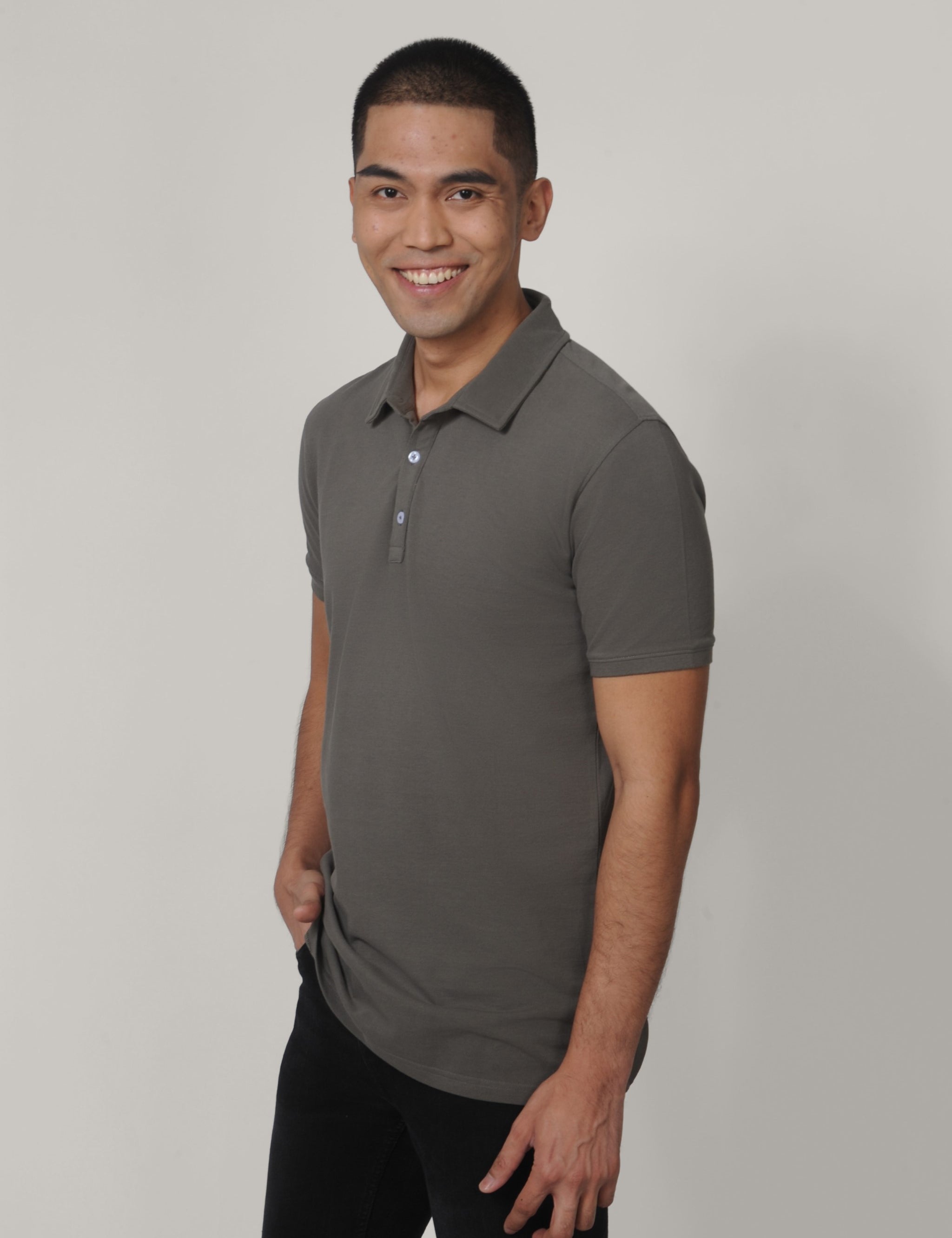 A tall and slim man in the studio standing in front of a light background. The smiling model is wearing an extra long slim dark grey polo shirt in a size medium. The tall dark grey polo shirt features a 3" longer body, 100% organic cotton, and is soft & preshrunk. The dark grey polo shirt is ideal for tall slim men 6'2"+.