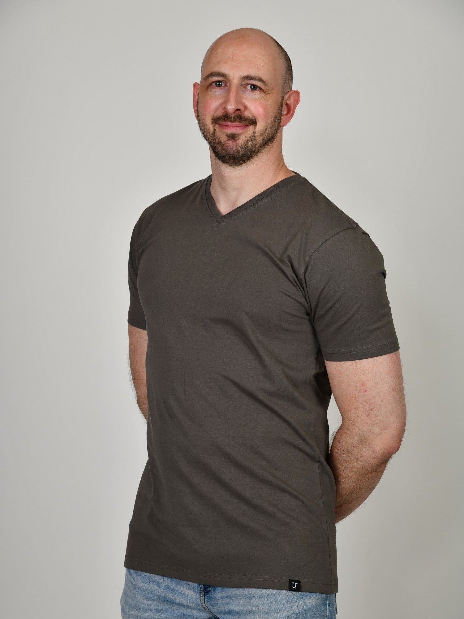 A tall and slim man in the studio standing in front of a light background with hands behind back. The smiling model is wearing an extra long slim dark grey v-neck t-shirt in a size XL. The tall dark grey v-neck t-shirt features a 3" longer body, 100% organic cotton, and is soft & preshrunk. The dark grey v-neck t-shirt is ideal for tall slim men 6'2"+.