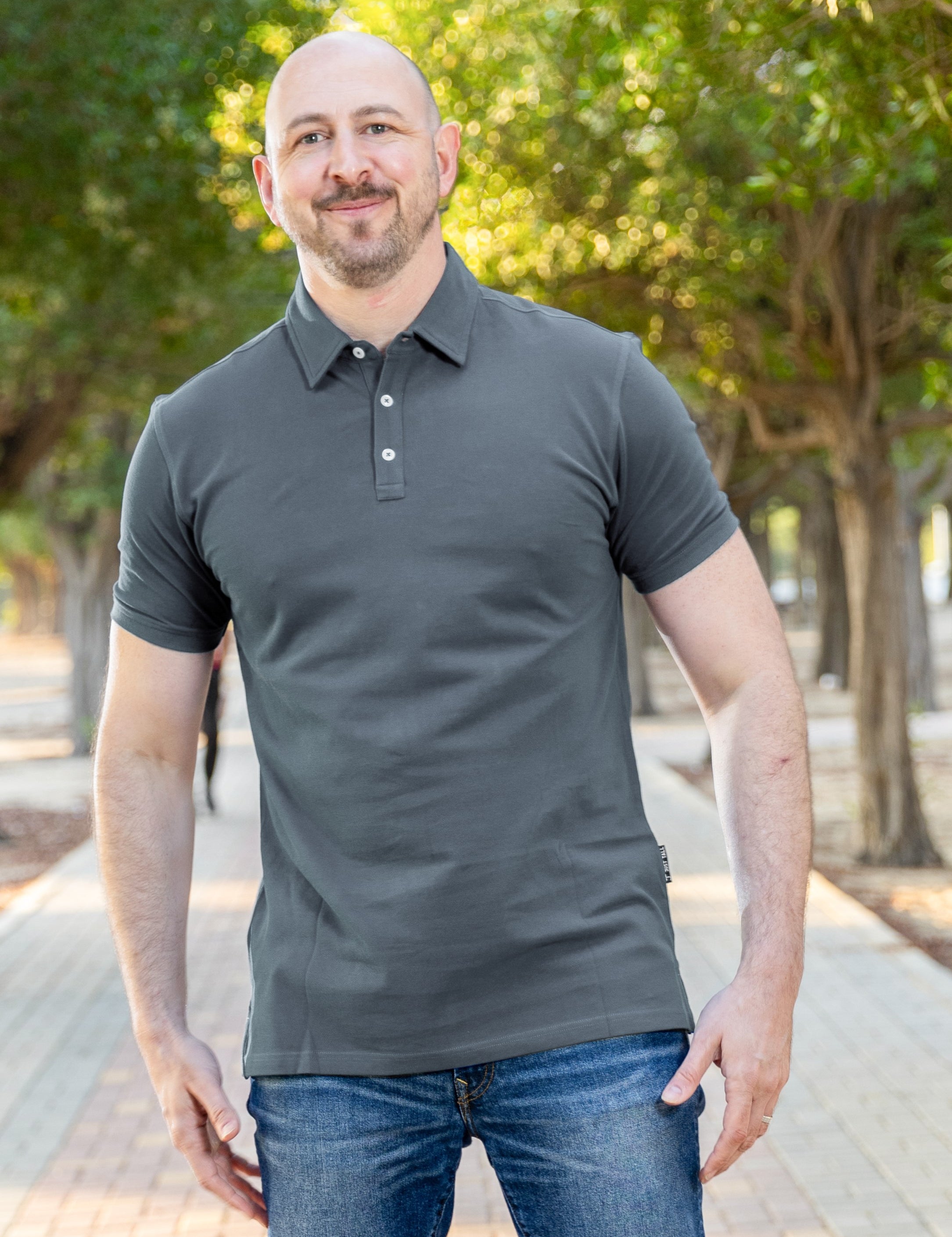 A tall and athletic man in a park standing on a path between trees. The smiling model is wearing an extra long slim dark slate polo shirt in a size XL. The tall dark slate polo shirt features a 3" longer body, 100% organic cotton, and is soft & preshrunk. The dark slate polo shirt is ideal for tall slim men 6'2"+. Buy 2 Save 10%, Buy 4 Save 15%, Buy 6 Save 20%.