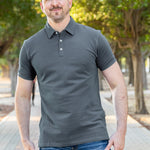 A tall and athletic man in a park standing on a path between trees. The smiling model is wearing an extra long slim dark slate polo shirt in a size XL. The tall dark slate polo shirt features a 3" longer body, 100% organic cotton, and is soft & preshrunk. The dark slate polo shirt is ideal for tall slim men 6'2"+. Buy 2 Save 10%, Buy 4 Save 15%, Buy 6 Save 20%.