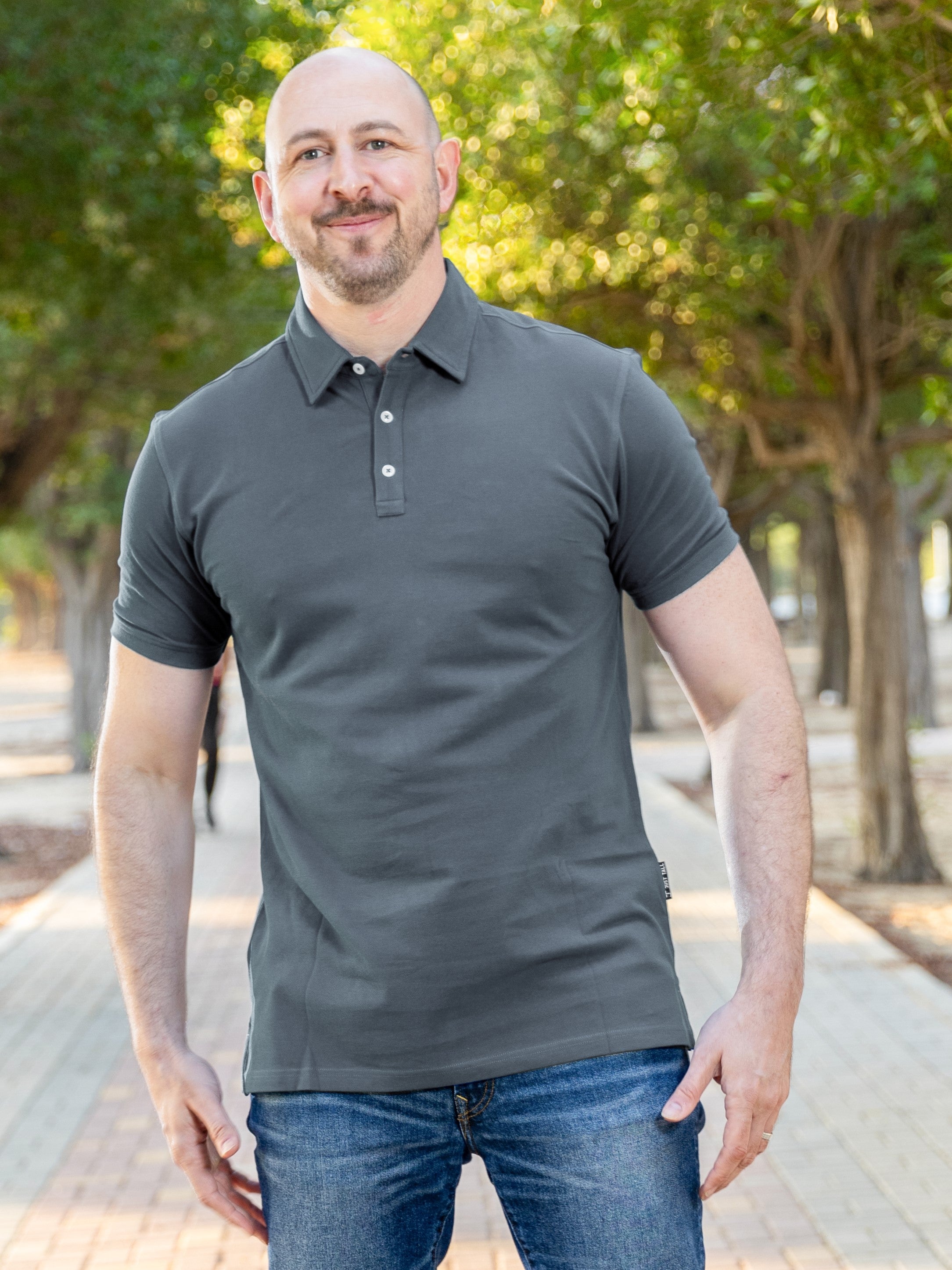 A tall and athletic man in a park standing on a path between trees. The smiling model is wearing an extra long slim dark slate polo shirt in a size XL. The tall dark slate polo shirt features a 3" longer body, 100% organic cotton, and is soft & preshrunk. The dark slate polo shirt is ideal for tall slim men 6'2"+. Buy 2 Save 10%, Buy 4 Save 15%, Buy 6 Save 20%.