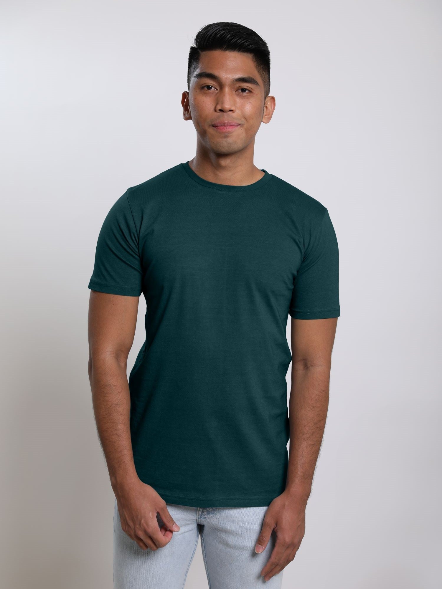 A tall and slim man in the studio standing in front of a light background with one hand in his pocket. The smiling model is wearing an extra long slim dark teal t-shirt in a size medium. The tall dark teal  t-shirt features a 3" longer body, 100% organic cotton, and is soft & preshrunk. The dark teal  t-shirt is ideal for tall slim men 6'2"+.