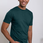 An upper body shot of a tall skinny guy wearing a dark teal medium tall t-shirt, hand in pocket.