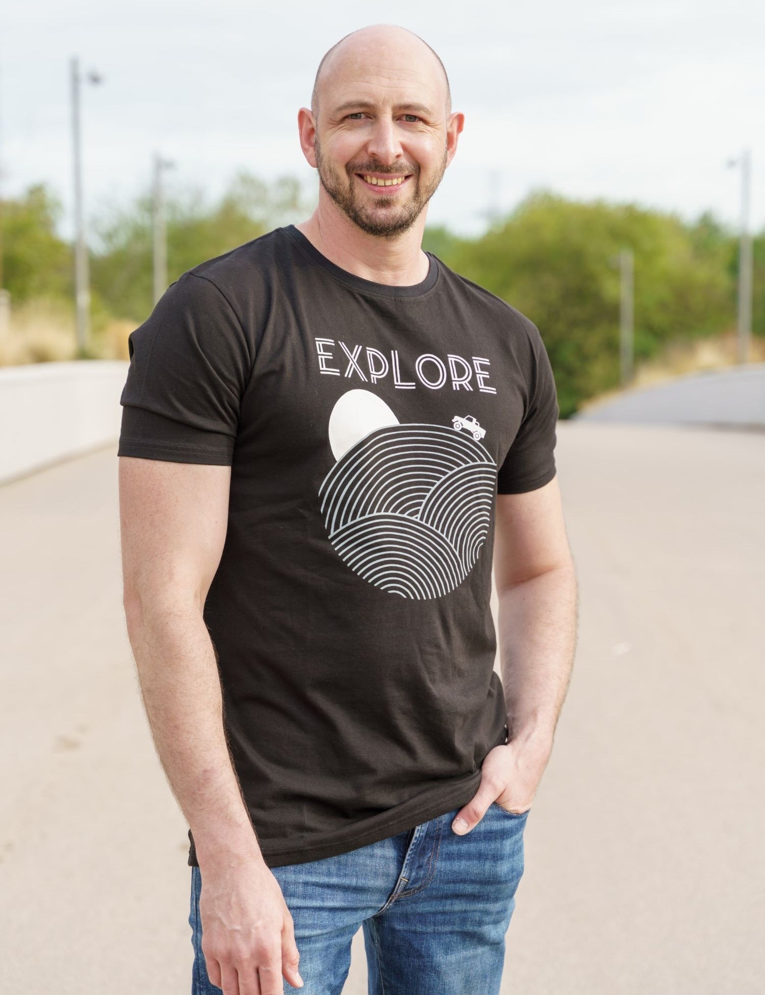 A tall and slim man standing in a park. The smiling model is wearing a black extra long graphic t-shirt with the text explore and a truck on the horizon. It features a 3" longer body, 100% organic cotton, and is soft & preshrunk. This extra long tall graphic tee is ideal for tall slim men 6'2"+.