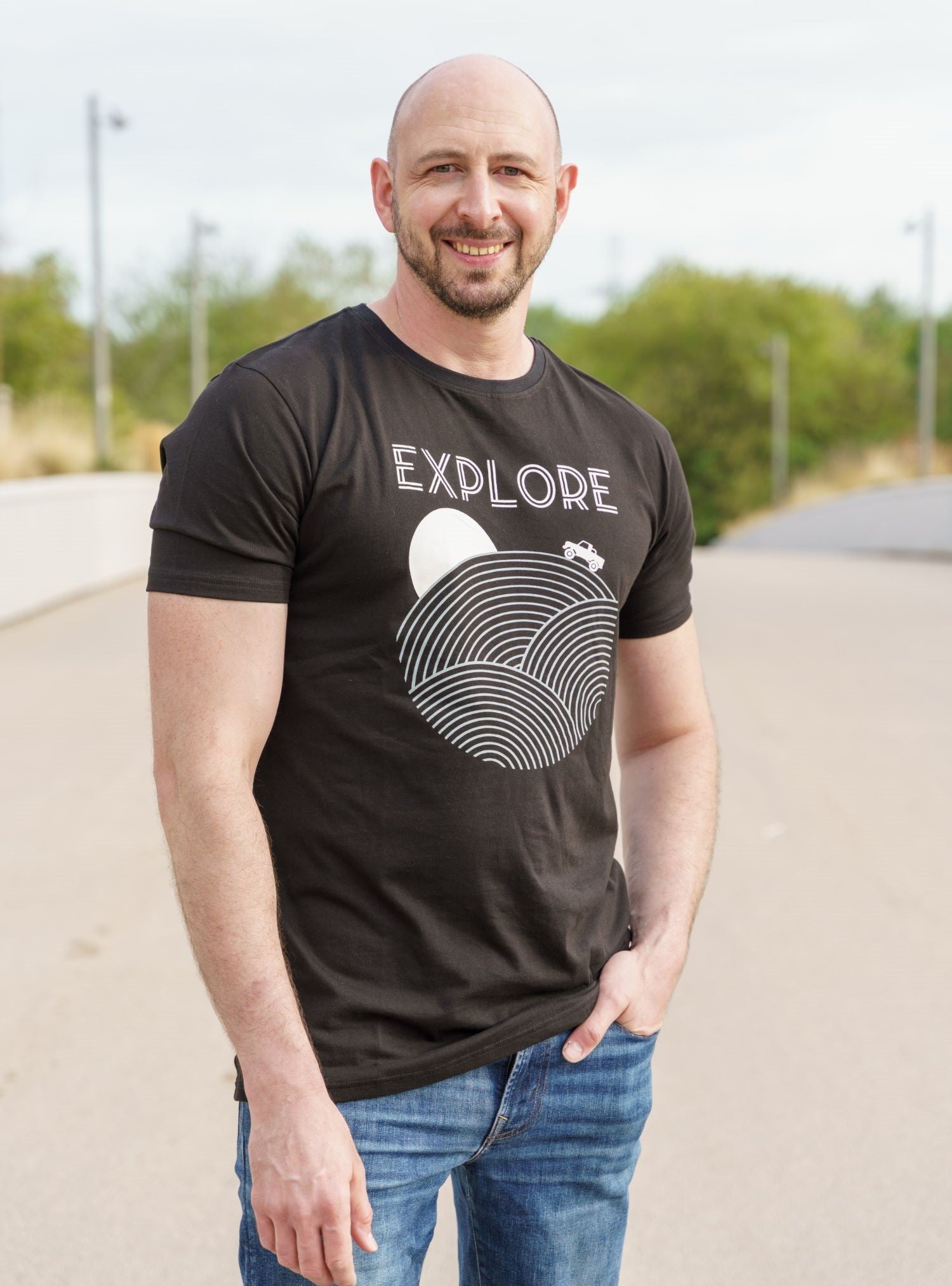 A tall and slim man standing in a park. The smiling model is wearing a black extra long graphic t-shirt with the text explore and a truck on the horizon. It features a 3" longer body, 100% organic cotton, and is soft & preshrunk. This extra long tall graphic tee is ideal for tall slim men 6'2"+.