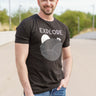 A tall and slim man standing in a park. The smiling model is wearing a black extra long graphic t-shirt with the text explore and a truck on the horizon. It features a 3" longer body, 100% organic cotton, and is soft & preshrunk. This extra long tall graphic tee is ideal for tall slim men 6'2"+.