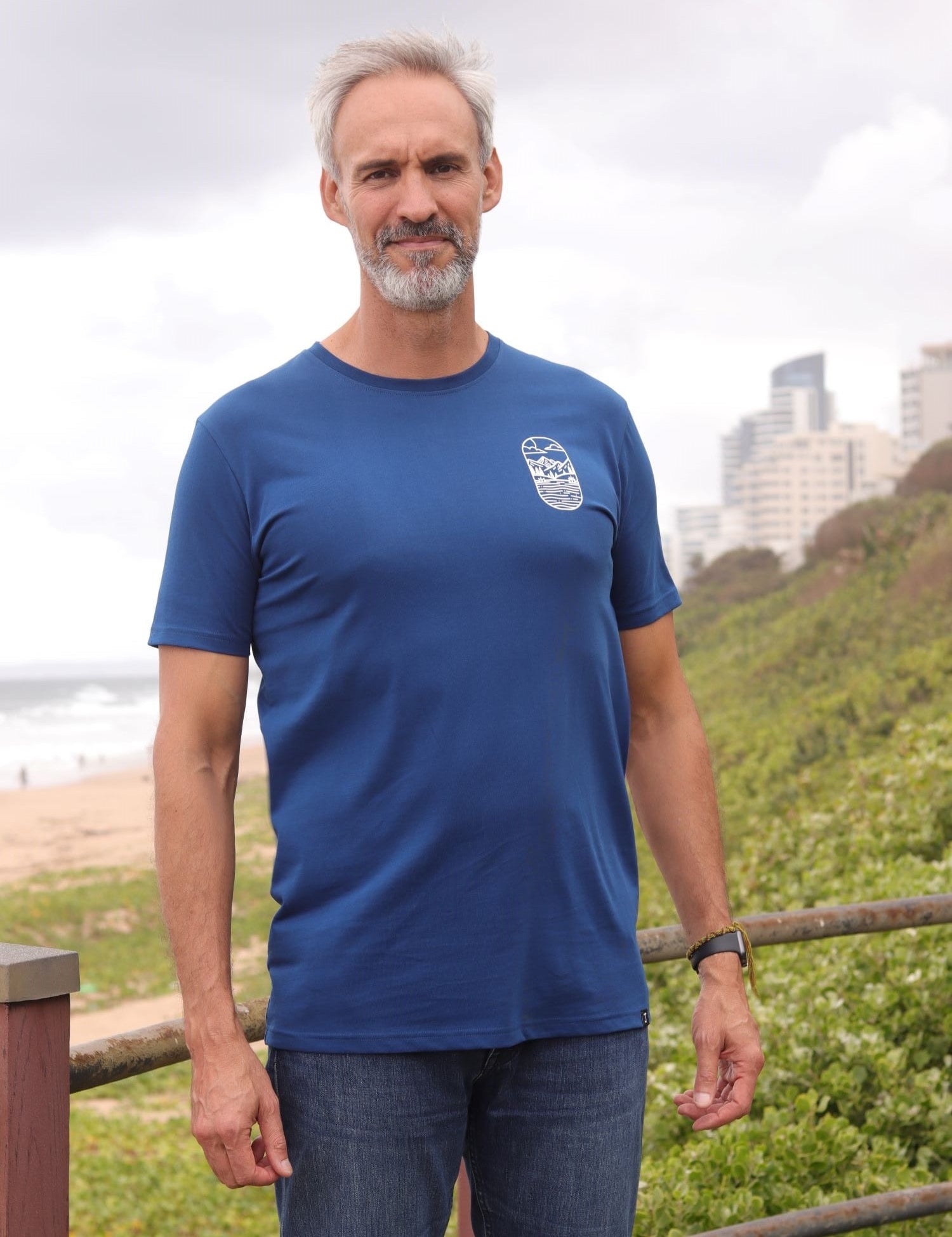 A tall and slim man standing by the sea. The smiling model is wearing a navy blue extra long graphic t-shirt with a minimal lake design. It features a 3" longer body, 100% organic cotton, and is soft & preshrunk. This extra long tall graphic tee is ideal for tall slim men 6'2"+