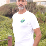 A tall and slim man standing by the sea. The smiling model is wearing a white extra long graphic t-shirt with a minimal landscape design. It features a 3" longer body, 100% organic cotton, and is soft & preshrunk. This extra long tall graphic tee is ideal for tall slim men 6'2"+.