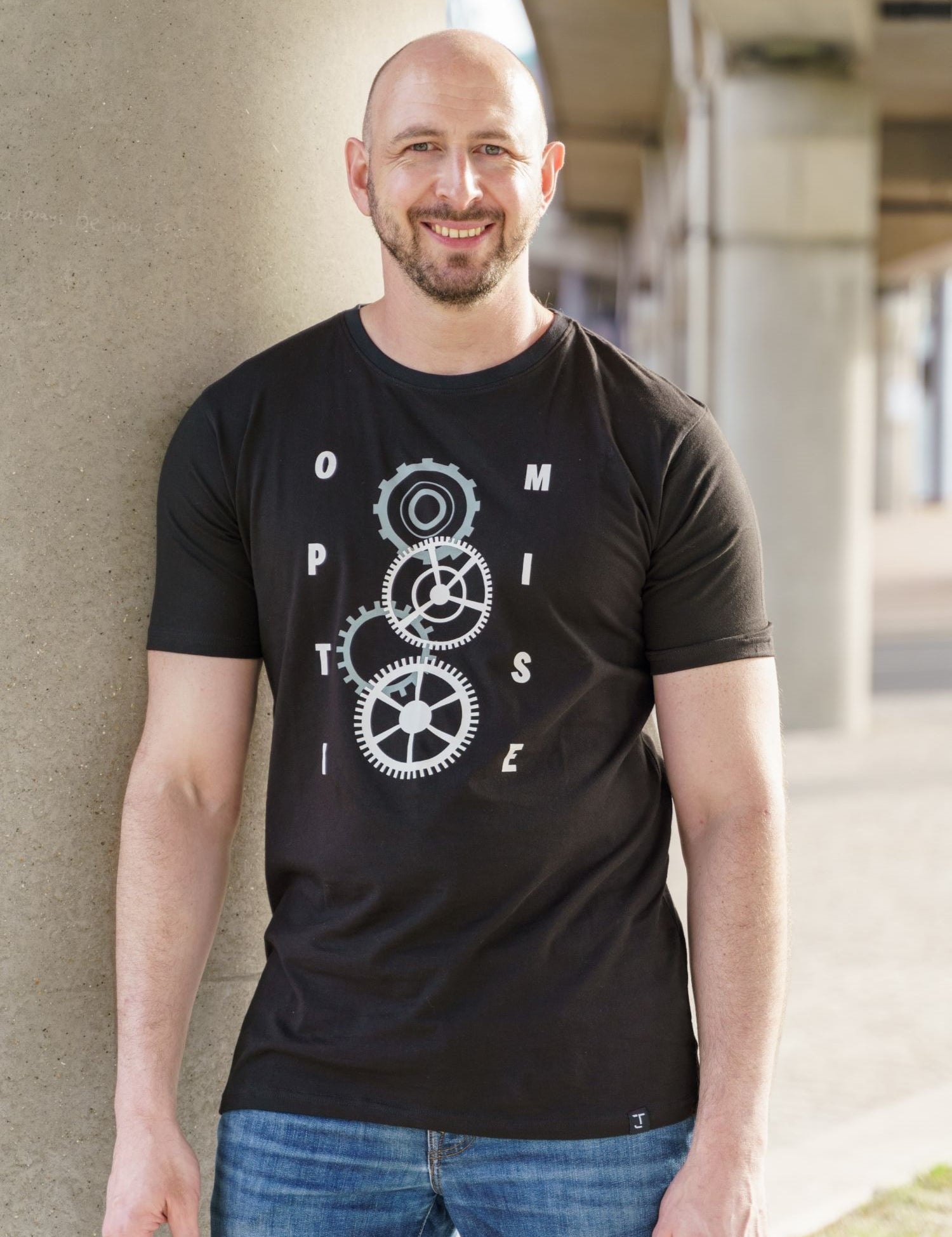 A tall and slim man standing outside under a bridge. The smiling model is wearing a black extra long graphic t-shirt with the text optimise and cogs in the middle. It features a 3" longer body, 100% organic cotton, and is soft & preshrunk. This extra long tall graphic tee is ideal for tall slim men 6'2"+.