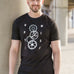 A tall and slim man standing outside under a bridge. The smiling model is wearing a black extra long graphic t-shirt with the text optimise and cogs in the middle. It features a 3" longer body, 100% organic cotton, and is soft & preshrunk. This extra long tall graphic tee is ideal for tall slim men 6'2"+.