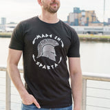 A tall and slim man standing by a canal. The smiling model is wearing a black extra long graphic t-shirt with the text made in sparta and a spartan helmet. It features a 3" longer body, 100% organic cotton, and is soft & preshrunk. This extra long tall graphic tee is ideal for tall slim men 6'2"+.