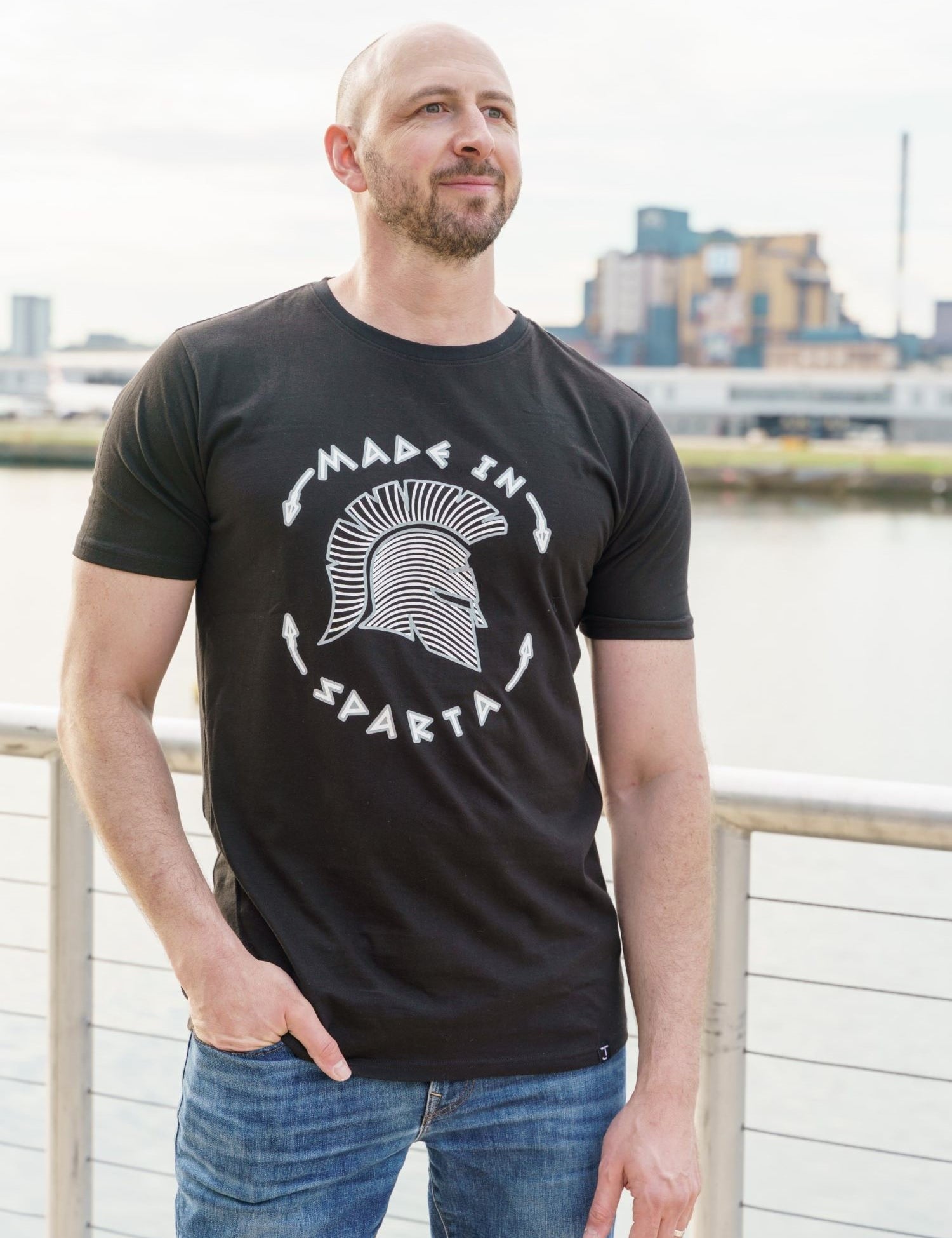 A tall and slim man standing by a canal. The smiling model is wearing a black extra long graphic t-shirt with the text made in sparta and a spartan helmet. It features a 3" longer body, 100% organic cotton, and is soft & preshrunk. This extra long tall graphic tee is ideal for tall slim men 6'2"+.
