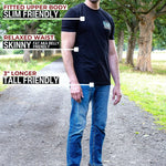 A tall man standing outdoors on a path wearing a black fitted t-shirt and blue jeans, with text callouts describing the shirt's fit: fitted upper body, relaxed waist, and 3" longer tall-friendly fit. Trees and grass are visible in the background.