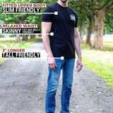 A tall man standing outdoors on a path wearing a black fitted t-shirt and blue jeans, with text callouts describing the shirt's fit: fitted upper body, relaxed waist, and 3" longer tall-friendly fit. Trees and grass are visible in the background.