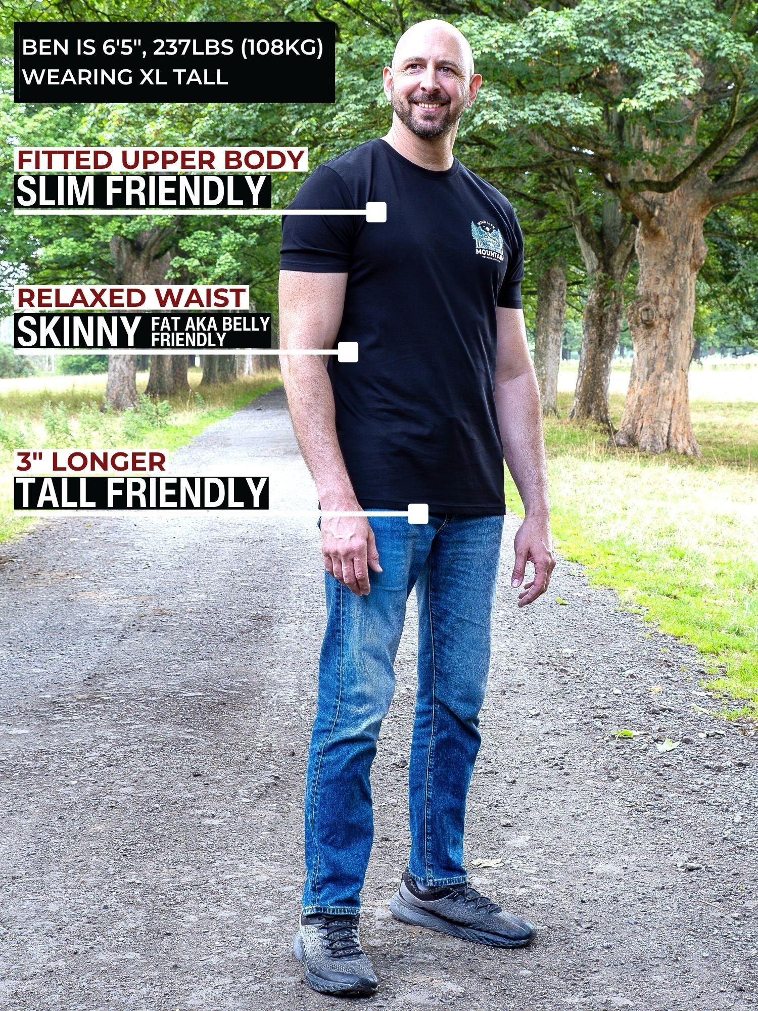 A tall man standing outdoors on a path wearing a black fitted t-shirt and blue jeans, with text callouts describing the shirt's fit: fitted upper body, relaxed waist, and 3" longer tall-friendly fit. Trees and grass are visible in the background.